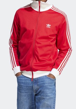 Osoba nosząca czerwoną kurtkę sportową Adidas z białymi paskami na rękawach i kołnierzu, zapiętą na zamek, z rękami w kieszeniach niebieskich dżinsów.