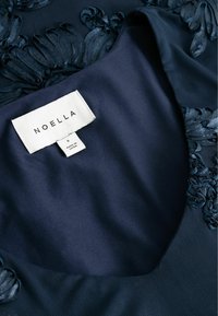 Tumši zils audums ar teksturētu ziedu izšuvumu apkaklē. Etiķete norāda "NOELLA," izmērs "S," un piezīme "Ražots Ķīnā."