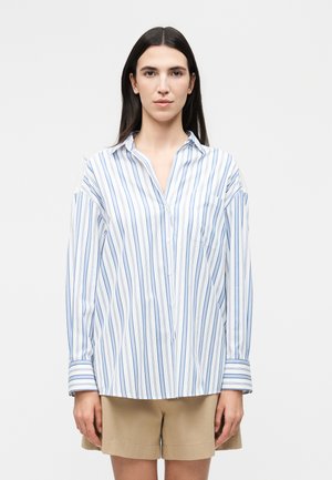 Kobieta o długich ciemnych włosach, ubrana w białą koszulę na guziki z niebieskimi pionowymi paskami i beżowe szorty, stojąca na jednolitym tle.