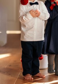 Valkoinen pitkähihainen paita, jossa on solmio, yhdistetty tummansinisiin housuihin. Lapsi seisoo paljain jaloin puulattialla, kädet ristissä.