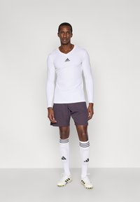 Fehér hosszú ujjú sportfelső fekete logóval, szürke rövidnadrág, fehér zokni fekete csíkokkal, valamint arany díszítésű futballcipő.