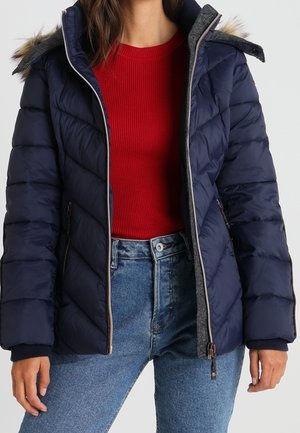 Femme portant une veste matelassée bleu marine avec capuche bordée de fourrure, un pull rouge côtelé et un jean bleu taille haute, debout devant un fond uni.