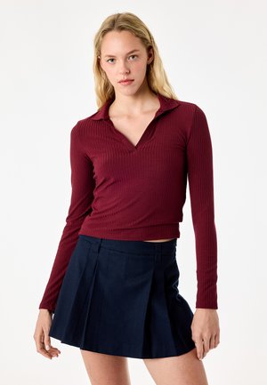 Giovane donna con capelli biondi che indossa una maglia bordeaux a costine manica lunga e una gonna plissettata blu navy su sfondo bianco.
