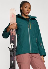 Teal waterdichte jas met een voorkleur rits en gele accenten, gecombineerd met lichtroze skibroeken. Model houdt bruine ski's met bindingen vast.