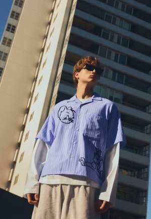 Mladý muž v slunečních brýlích, oblečený do pruhovaného trička s krátkými rukávy s kreslenými motivy přes dlouhé bílé rukávy, stojící před vysokými městskými budovami.