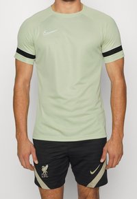 Hombre con camiseta deportiva Nike verde claro y pantalones cortos negros del Liverpool FC, con logotipos de Nike y acentos beige, de pie frente a un fondo liso.