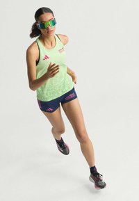 Svijetlozeleni top bez rukava s grafičkim tekstom, mornarske kratke hlače s ružičastim naglascima, i višebojne reflektirajuće sunčane naočale. Pozicija trčanja, atletske cipele.