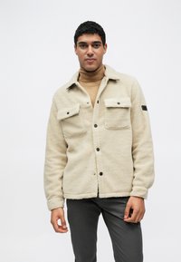 Μπεζ φλις μπουφάν με μαύρα κουμπιά, δύο τσέπες στο στήθος και λείο υφή. Φορεμένο πάνω από ένα ταμπά ζιβάγκο και γκρι παντελόνια.