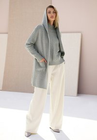 Grijze gebreide cardigan met een capuchon, over een getextureerde grijze trui, gecombineerd met crème wijde pijpen broek en lichtgrijze puntige schoenen.