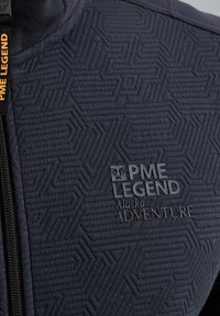 Section de veste matelassée foncée avec motif géométrique, fermeture éclair et logo "PME Legend Alaska Adventure" en gris.