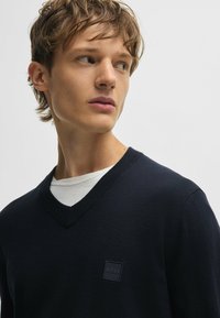 Mörkblå stickad tröja med V-hals, med en strukturerad krage och en liten svart fyrkantig logotyp på bröstet. Under tröjan har personen på sig en vit skjorta.