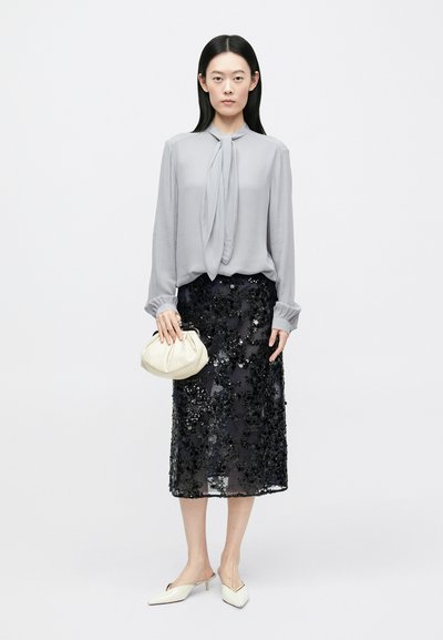 Kobieta w jasnoszarej bluzce z wiązaniem pod szyją, czarna spódnica midi z cekinami, białe szpiczaste buty, trzymająca małą białą torbę kopertówkę.