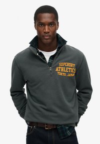 Sudadera gris con cremallera de un cuarto que presenta el texto amarillo "Superdry Athletics Tokyo", sobre una camisa de cuadros azul, con una textura suave.