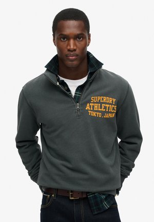 Sudadera gris con cremallera de un cuarto que presenta el texto amarillo "Superdry Athletics Tokyo", sobre una camisa de cuadros azul, con una textura suave.