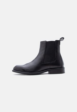 Botte Chelsea en cuir noir avec panneau latéral élastique, languette de traction au talon, petit talon bloc et bout arrondi lisse sur fond blanc.