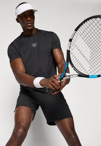 Melnā sporta krekls ar logo uz krūtīm, apvienots ar melniem šortiem. Tenisa spēlētājs tur racket, valkājot baltu vizieri un plaukstas siksnu.