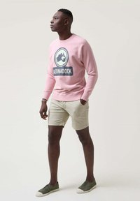 Sudadera rosa claro con un gran logo gráfico negro, combinada con pantalones cortos beige y zapatillas verdes. Textura suave, ajuste casual.