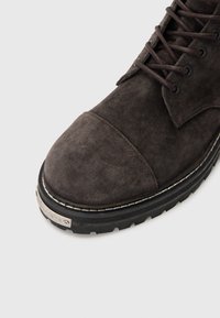 Sive čizme od suede materijala s zaobljenim vrhom, vezanjem sprijeda i teksturiranom gumenu potplatom. Imaju metalni detalj na peti.