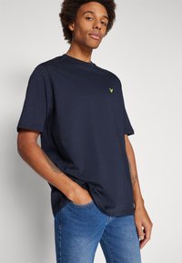 Marineblauwe katoenen t-shirt met een ronde hals, korte mouwen en een klein geel logo op de borst. Draag met blauwe denimjeans.