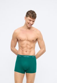 Žalios medvilninės boxer tipo apatines kelnaites su juodu "Calvin Klein" logotipu juosmenyje, pasižyminčios prigludusia forma ir lygiu paviršiumi. Modelis stovi atsisukęs į priekį, šypsosi.