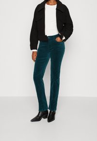Teal kordfluweelbyxor med slim fit, med framfickor och en strukturerad yta, matchade med en svart kort jacka och spetsiga svarta stövlar.