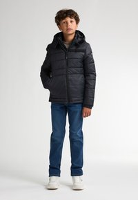 Zwarte pufferjack met een capuchon, voorzien van een rits aan de voorkant, zijzakken en geribbelde manchetten, gecombineerd met blauwe denim jeans en witte sneakers.