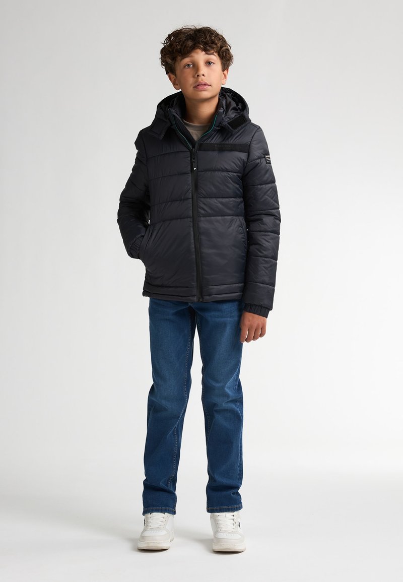 Zwarte pufferjack met een capuchon, voorzien van een rits aan de voorkant, zijzakken en geribbelde manchetten, gecombineerd met blauwe denim jeans en witte sneakers.