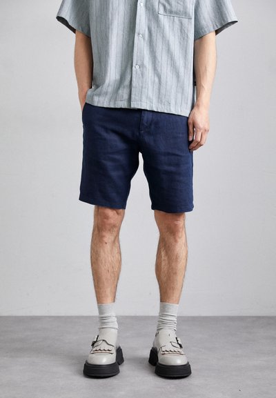Pantalones cortos azul marino de corte holgado, hechos de tela ligera, combinados con una camisa de rayas gris y zapatos grises distintivos con suelas negras.