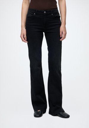 Personne portant un jean bootcut taille haute noir, des chaussures noires et un haut sans manches marron foncé, debout devant un fond clair uni.