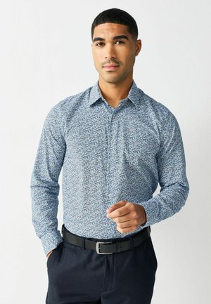 Langärmliges Hemd mit Knopfleiste in Blau mit floralem Muster, ausgestattet mit einem Umlegekragen und einer Brusttasche, kombiniert mit dunklen Hosen und einem schwarzen Gürtel.