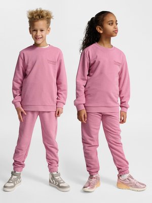 To børn står og har matchende lyserøde sweatshirts og bukser på, det ene smiler fremad, det andet kigger til siden.