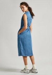 Robe midi en denim bleu clair avec une taille ceinturée, sans manches, poches latérales et détails de couture. Mannequin portant des baskets grises.