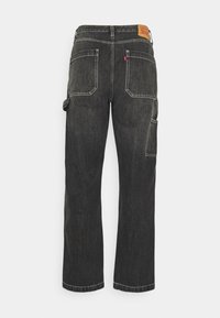 Pantalon de travail en denim noir vu de dos avec deux grandes poches, une boucle pour outils sur la jambe droite et un patch de marque en cuir marron à la ceinture.