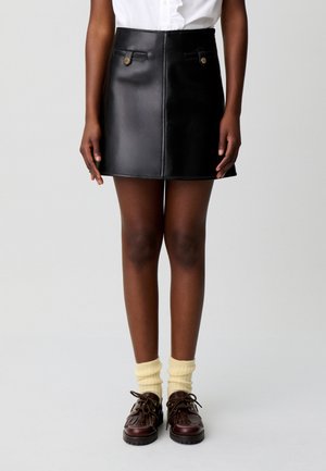 Personne portant une mini-jupe en cuir noire avec des poches avant, des chaussettes crème et des chaussures marron foncé à lacets, debout devant un fond uni.