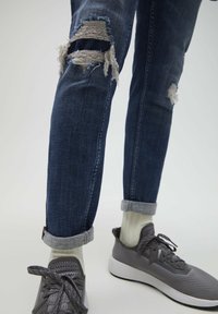 Ben med mörkblå slitna jeans som är uppvikta vid fållen, vita ribbade strumpor och grå mesh-atletiska skor på en ljus bakgrund.