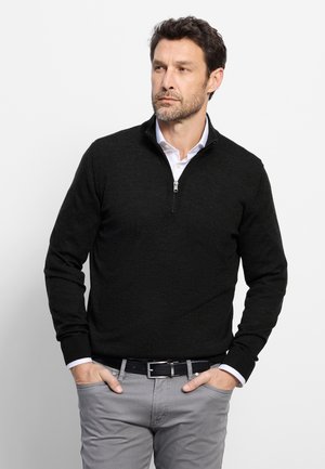 Uomo che indossa un maglione nero con cerniera sopra una camicia bianca con pantaloni grigi, mani in tasca, guarda di lato su uno sfondo bianco uniforme.