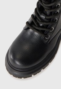 Bota de cuero negro con suela gruesa, ojales de metal y cordones redondos negros, mostrada en primer plano sobre un fondo blanco.