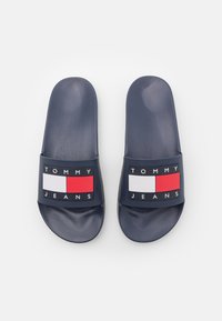 Tumesinised libisemise sandaalid, millel on lai rihm ja millel on "TOMMY JEANS" tekst ning ristkülikukujuline logo punase ja valgega. Sile tekstuur, madal profiil.