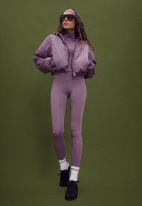 Lila Puff-Jacke mit hohem Kragen, passenden Leggings und schwarzen Sneakers. Ausgestattet mit großen Sonnenbrillen und kontrastierenden weißen und lila Socken.
