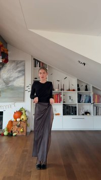 Μακριά, σκούρα καφέ σατέν φούστα με πλαϊνό άνοιγμα, συνδυασμένη με εφαρμοστό μαύρο τοπ με μακριά μανίκια. Ξύλινο πάτωμα και διακοσμημένα ράφια στο φόντο.