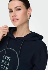Felpa con cappuccio blu navy con una grafica circolare "Copenaghen". Presenta un cappuccio con cordino e una texture morbida, mostrando un design informale.