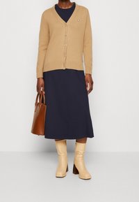 Kamelstrikket cardigan med V-hals, seks knapper, marineblå kjole, brun håndtaske og beige støvler med hæl, firkantet tå og tekstureret finish.