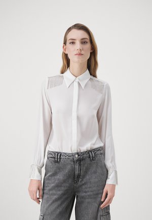 Blusa bianca a maniche lunghe con colletto, spalle decorate e rifiniture ai polsini. Tessuto setoso, leggero e dal design aderente.