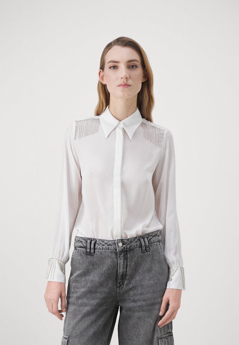 Blusa bianca a maniche lunghe con colletto, spalle decorate e rifiniture ai polsini. Tessuto setoso, leggero e dal design aderente.
