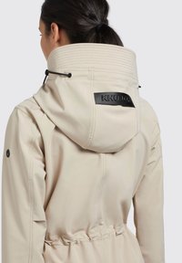 Beige vattentålig jacka med hög krage, svart logotyp-patch, dragsko i huvan och elastisk midja. Har en slät, strukturerad tyg.