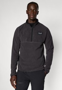 Pulover din fleece de culoare gri închis, cu guler înalt, închidere cu fermoar pe jumătate și etichetă pe piept. Textură moale și mâneci lungi.