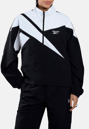 Frau trägt eine schwarz-weiße Reebok-Jacke mit Reißverschluss und geometrischem Design sowie passende schwarze Hosen, steht mit einer Hand in der Tasche.