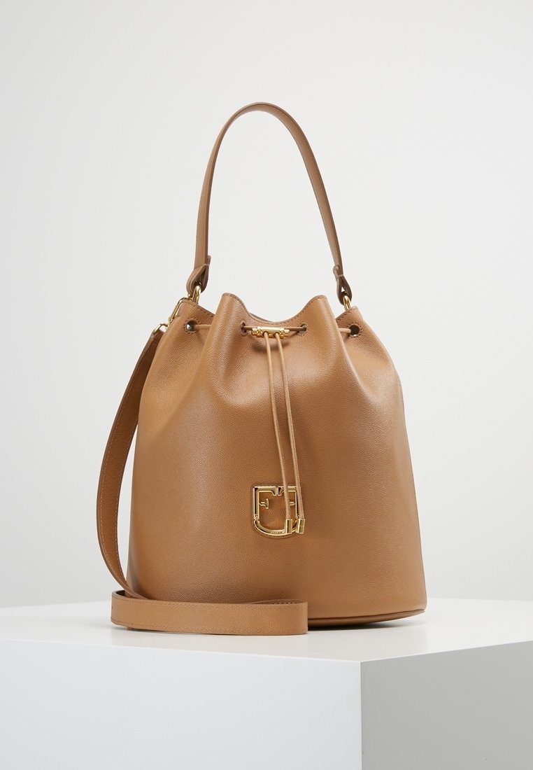 Sac seau en cuir camel avec fermeture à cordon dorée, poignée supérieure, bandoulière amovible et charm logo doré décoratif.