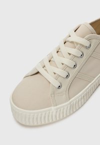 Beige kangaskengät, joissa on pyöreä kärki, valkoinen kumipohja ja erottuvat nauhaketjut. Varusteena metalliset silmukat ja tekstuuria pohjan varrella.
