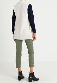 Stickad ärmlös väst i ljusbeige, kombinerad med en marinblå långärmad skjorta och olivgröna byxor; svarta ankelboots fulländar outfiten.